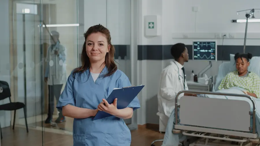 A nurse in blue scrubs holding a clipboard on a hospital ward